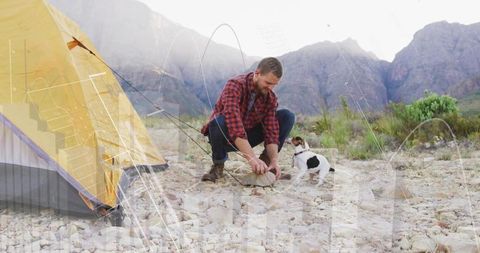 Kneeling hiker tying boots at rocky mountain campsite with yellow tent and small white dog