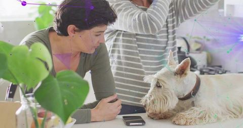 Woman leaning and watching white terrier on kitchen counter with smartphone and monstera