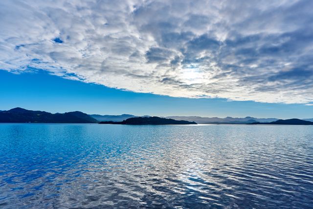 This tranquil scene shows a calm sea with gentle waves and reflections, surrounded by scenic mountains. The sky above is filled with striking clouds that add depth to the image. Ideal for use in websites and prints that promote relaxation, nature, travel, tourism, wellness retreats, or backgrounds for motivational quotes.