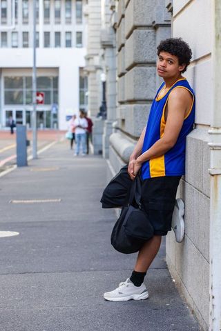Urban basketball player leaning on stone facade carrying duffel bag in blue yellow jersey