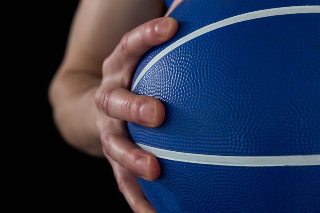 This image shows a close-up of a player's hand gripping a blue basketball against a black background. Ideal for use in sports-related content, advertisements for basketball equipment, athletic training materials, and motivational posters.