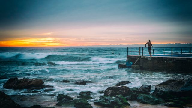 This image captures a man standing on a pier as he enjoys the sunrise over the ocean waves. The scene is set with the rocky coastline in the foreground and a cloudy sky creating a tranquil atmosphere. Perfect for use in travel blogs, relaxation and wellness articles, coastal living promotions, or nature photography collections.