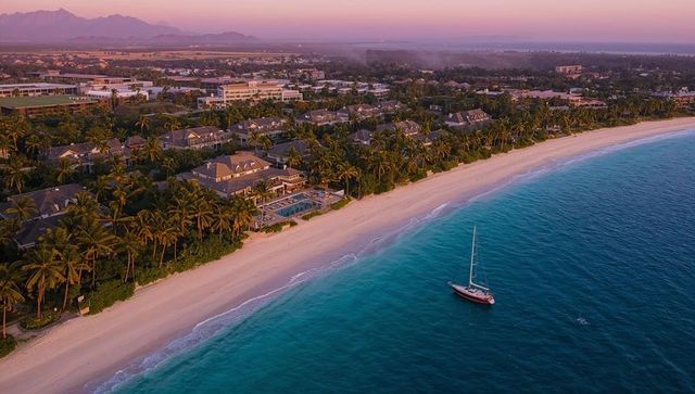 Turquoise oceanfront resort, palm lined white sand beach curving along shore with sailboat