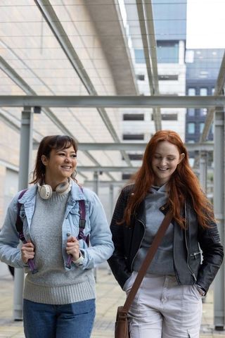 Multicultural young women walking on urban campus under glass canopy, smiling and chatting
