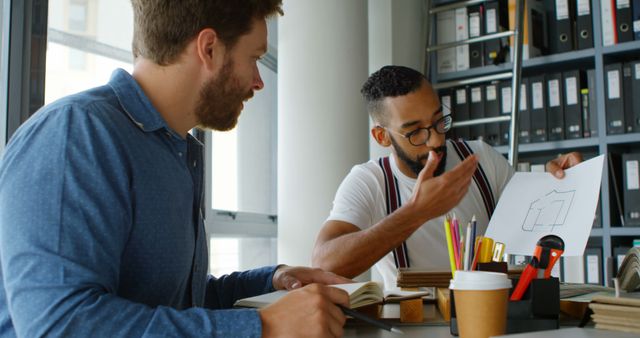 Bearded man wearing denim shirt discussing apartment floor plan with diverse coworker holding paperwork in bright modern office. Ideal for use in articles about team collaboration, architectural planning, workplace diversity, interior design projects, professional teamwork, and productivity in creative spaces.