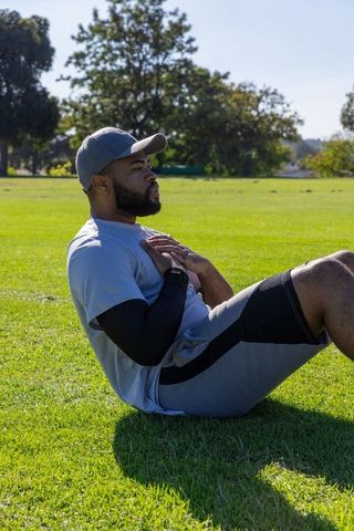 African american man performing sit-up in park wearing baseball cap and fitness watch