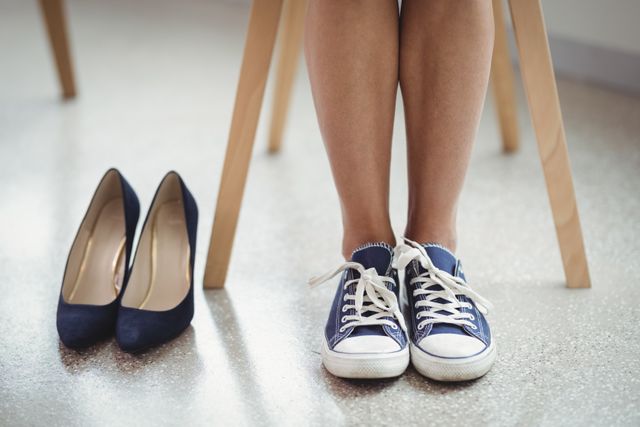 Woman's legs in canvas shoes with high heels placed nearby on office floor. This image can be used to depict themes of comfort versus formality, work-life balance, or personal choice in professional settings.
