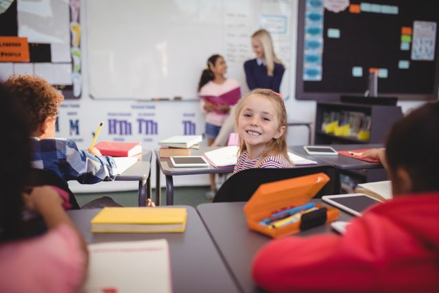 This image shows a cheerful schoolgirl sitting at her desk in a classroom, with a teacher and other students in the background. Ideal for educational materials, school advertisements, and articles about primary education and classroom environments.