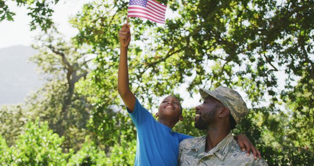 Happy african american soldier father carrying son waving flag in garden. Military service, fatherhood, patriotism, family and togetherness.