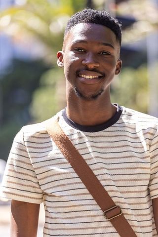 Young man smiling outdoors wearing striped t-shirt and canvas crossbody strap, sunlit portrait