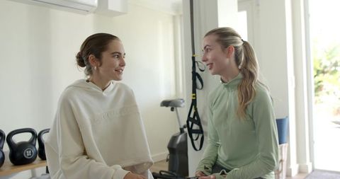 Female friends chatting in bright home gym holding phone after workout session