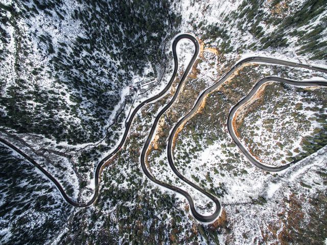 This image captures a winding mountain road covered in snow viewed from above. The curving path meanders through the snowy landscape, showcasing the beauty of nature during winter. Dense forest surrounds the road, with pine trees dotting the rugged terrain. Ideal for use in travel and adventure articles or promotions, winter landscape advertisements, and outdoor lifestyle blogs.