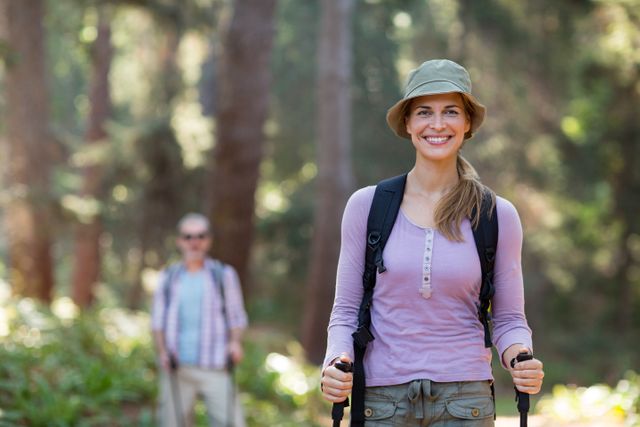 Smiling woman hiking with trekking poles in a lush forest. She is wearing a hat and backpack, enjoying the outdoor adventure. Ideal for promoting healthy lifestyles, outdoor activities, travel, and nature exploration.