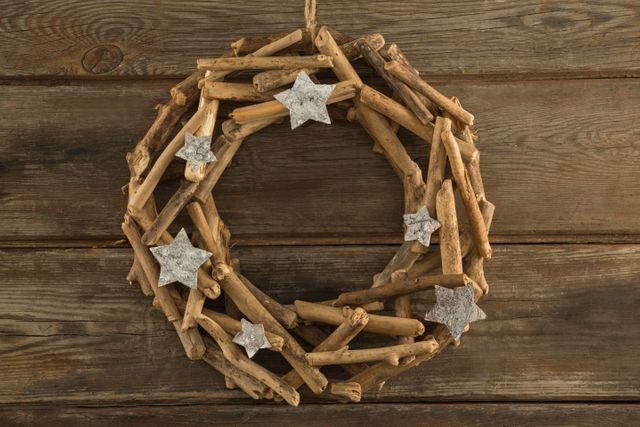 Rustic Christmas wreath made of natural wooden sticks and adorned with silver stars, hanging on a wooden plank background. Ideal for holiday-themed promotions, home decor inspiration, festive greeting cards, and seasonal blog posts.