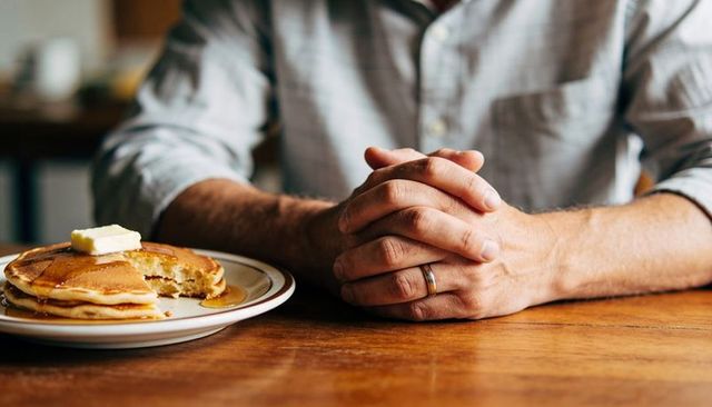Man resting folded hands beside pancakes with butter and syrup on wooden breakfast table
