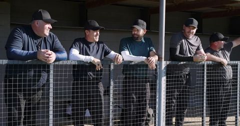 Baseball coaches leaning on dugout railing watching players and chatting in sunlit field