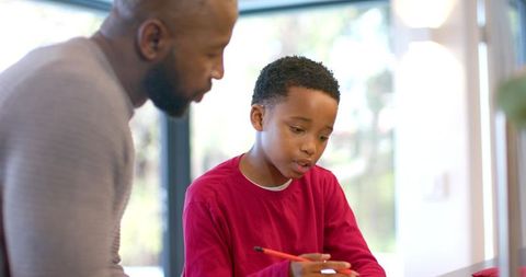 African american father guiding son writing homework at bright home desk, tutoring moment