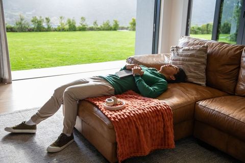 Relaxing young man lounging on leather sectional by open glass doors with green lawn and cozy throw