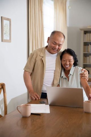 Chinese senior couple smiling while using laptop at cozy table for online banking and video calls
