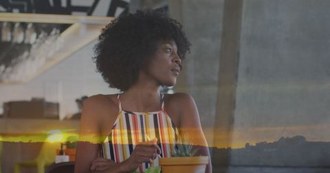 This striking image features a contemplative African American woman with curly hair, seated at a table with a salad, overlaid with a serene sunset landscape. The juxtaposition of indoor dining against the outdoor calm evokes themes of reflection and serenity. Ideal for lifestyle blogs, adverts focusing on mindfulness and relaxation, or dining experiences that offer tranquility and escape.