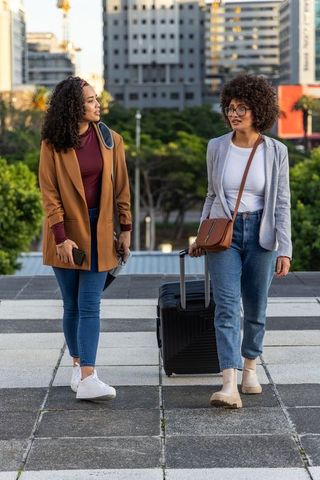 Young women walking with suitcase in urban plaza conversing during city travel