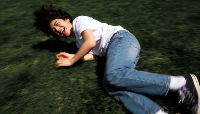 Teen rolling and laughing on lawn wearing white tee and blue jeans, candid motion blur