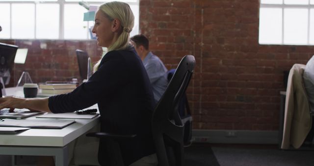 Businesswoman sitting at desk, typing on computer in a modern, open-plan office with exposed brick walls and large windows. Suitable for concepts related to professional work environment, corporate culture, technology use in business, and modern workplace settings.