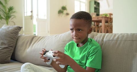 African american boy concentrating playing console game holding white controller on sofa