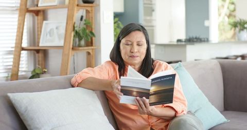 Mature asian woman sitting on sofa reading paperback endless sunsets in sunlit living room