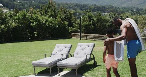 Father guiding son on sunny lawn by chaise lounges, summer backyard bonding
