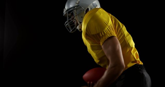 This is an image of a football player wearing a yellow jersey and helmet, captured holding a football against a black background. The image showcases athleticism, strength, and focus, making it suitable for use in sports advertising, fitness-related content, and motivational posters. It can also be used in articles and promotional materials related to football and team sports.