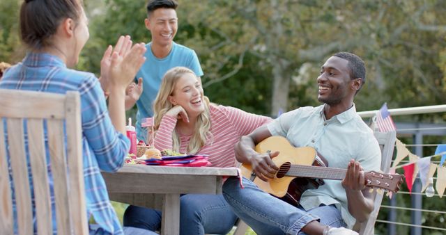 Friends sitting outside, enjoying a picnic with smiles and laughter. One person playing a guitar while others listen and applaud. Festive atmosphere with food, drinks, and decorative flags. Perfect for ads about social gatherings, friendship, summer events, and casual get-togethers.