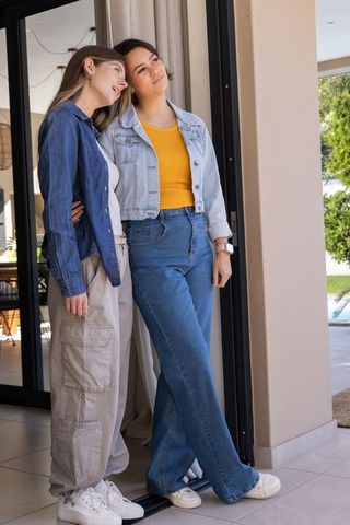 Sunlit diverse female couple standing at sliding door leaning together in denim and jeans