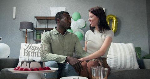 Couple holding hands on sofa celebrating birthday with cake, balloons and cozy living room