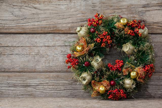 This image features a beautifully crafted Christmas wreath adorned with red berries, golden ornaments, and green foliage, leaning against a wooden background. Ideal for use in holiday greeting cards, festive advertisements, seasonal blog posts, and home decor inspiration.