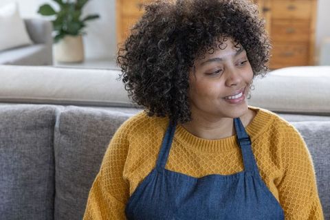 African american woman wearing denim apron and mustard sweater smiling and relaxing on gray sofa