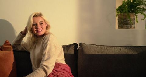 Young blonde woman relaxing on cozy dark gray sofa with knit blanket and warm sunlight