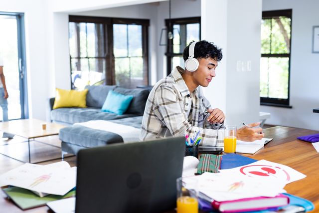 Biracial teenage boy with headphones and laptop doing homework at home. E-learning, home schooling and lifestyle concept.
