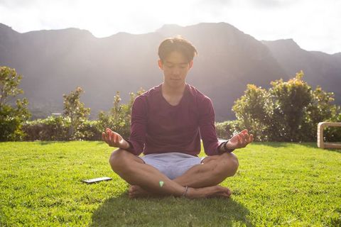 Sunlit asian man meditating cross-legged on lawn with mountain backdrop and smartphone