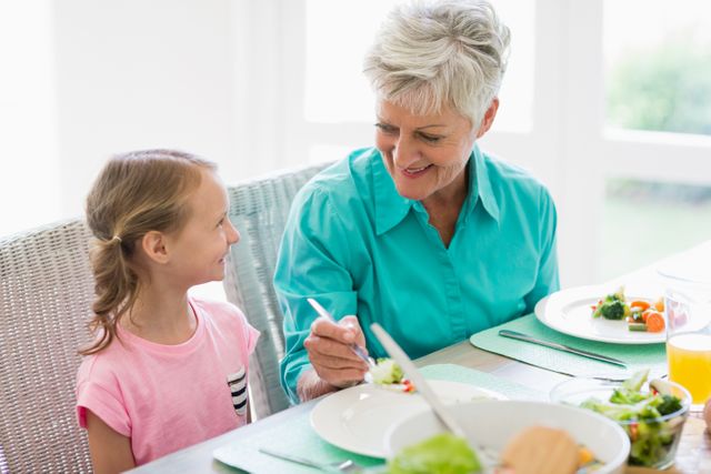 Grandmother serving food to her granddaughter at home, both smiling and enjoying their time together. Ideal for use in family-oriented content, healthy eating promotions, and advertisements focusing on intergenerational relationships and home life.
