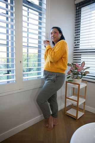 Mature woman standing by sunlit shutters holding mug in cozy mustard sweater morning calm