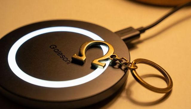 Black matte wireless charger featuring glowing white ring and brass omega keyring on warm desk