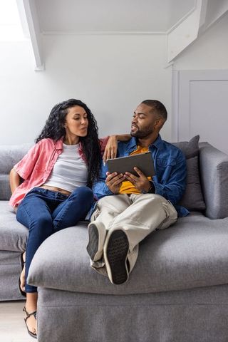 Couple relaxing on gray sectional sofa sharing tablet, african american man and partner
