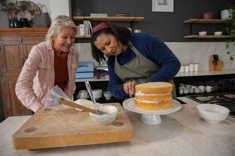 Two women assembling and smoothing frosting on layered sponge cake in cozy home kitchen