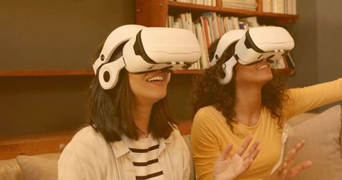 Two women experiencing virtual reality together on cozy sofa with white VR headsets