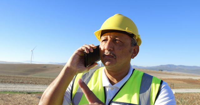 Male construction worker wearing a hard hat and safety vest is talking on a smartphone while pointing. Wind turbines are visible in the background under a clear blue sky. Can be used for themes related to construction, engineering, renewable energy, communication in remote locations, safety on job sites, and sustainable development.