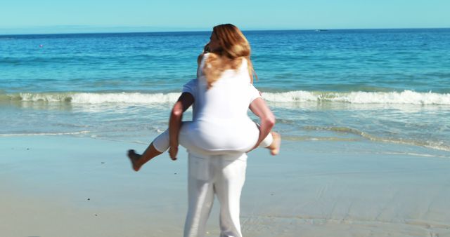 Couple enjoying a sunny day at the beach with piggyback ride in white clothes by ocean waves. This joyful, carefree scene can be used in campaigns promoting vacation destinations, romantic getaways, summer apparel, or beach resorts, as well as in articles or brochures that celebrate love and relationships.