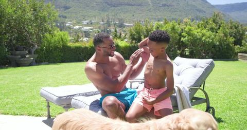 African american father playing with teenage son on sunny backyard lounge with family dog