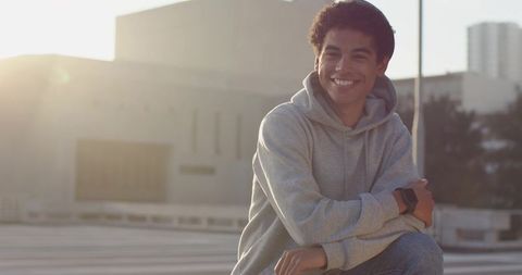 Smiling young man in gray hoodie checking smartwatch on rooftop at sunset with city skyline