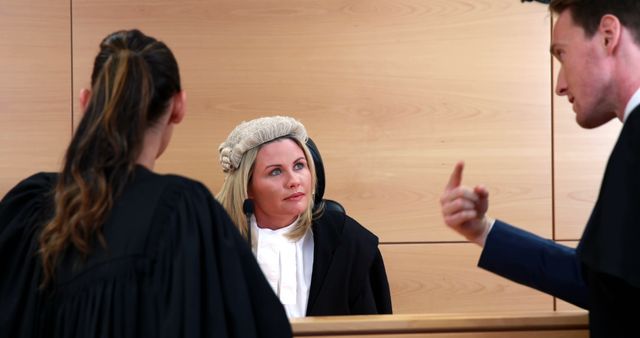 A Caucasian female judge attentively listens to a Caucasian male lawyer presenting his case in a courtroom, with a female lawyer of the same ethnicity observing the interaction. The image captures a critical moment in legal proceedings, emphasizing the gravity of judicial decision-making.